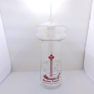 May include: A clear glass drinking glass shaped like the Skylon Tower in Niagara Falls, Canada. The glass has a red and white graphic of the tower with the text "Skylon Tower Niagara Falls, Canada".