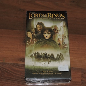 Peut inclure: Un boîtier DVD pour le film "Le Seigneur des anneaux : La Communauté de l'anneau". Le boîtier présente une scène du film avec plusieurs personnages, dont Frodon Sacquet, Gandalf et Aragorn. Le texte "REAL MAGIC" et "ONE OF THE BEST FILMS OF THE YEAR." est imprimé sur le boîtier.