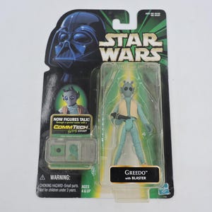 Könnte beinhalten: Star Wars Actionfigur von Greedo mit Blaster in Originalverpackung. Die Figur ist hellblau mit einer beigen Weste. Die Verpackung zeigt Darth Vader und das Star Wars-Logo. Die Verpackung enthält einen CommTech-Chip.