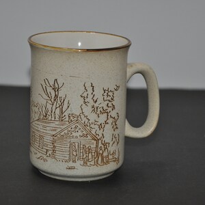 Könnte beinhalten: Eine beige Keramiktasse mit einer braunen Illustration einer Hütte im Wald. Die Tasse hat einen braunen Rand und einen braunen Henkel.