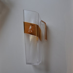 May include: A clear plastic decanter with a gold-coloured metal band and a walking man logo. The decanter has a handle and a textured surface.