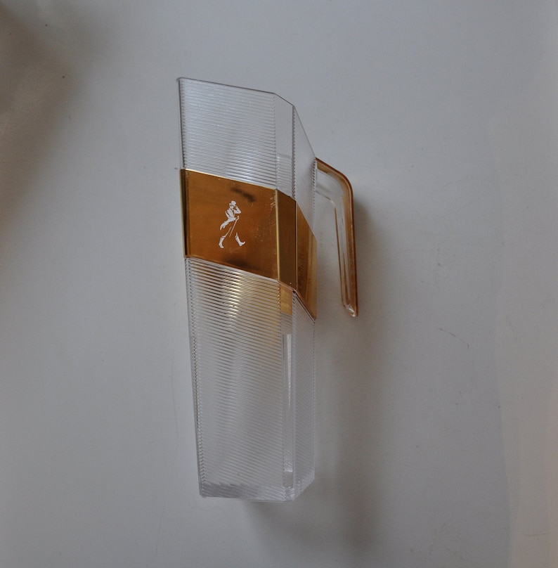 May include: A clear plastic decanter with a gold-colored metal band and a walking man logo. The decanter has a handle and a textured surface.