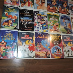 Pu&ograve; includere: Una collezione di videocassette VHS Disney vintage con classici animati come "Red e Toby nemiciamici", "La carica dei 101" e "Aladdin". Le cassette presentano illustrazioni colorate e sono disposte su una superficie di legno.