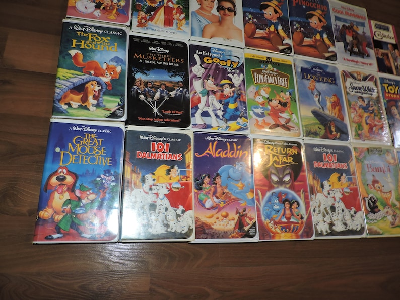 Pu&ograve; includere: Una collezione di videocassette VHS Disney vintage con classici animati come "Red e Toby nemiciamici", "La carica dei 101" e "Aladdin". Le cassette presentano illustrazioni colorate e sono disposte su una superficie di legno.