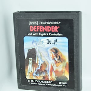 Könnte beinhalten: Eine schwarze Sears Tele-Games-Cartridge für das Videospiel Defender. Die Cartridge zeigt ein Bild einer Frau und eines Kindes vor einem Gebäude mit dem Text "DEFENDER" und "Mit Joystick-Controllern verwenden".