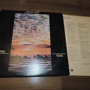 Könnte beinhalten: Ein Vintage-Vinyl-Schallplattenalbum von Split Enz mit dem Titel "Time and Tide". Das Albumcover zeigt einen Sonnenuntergang über einem Gewässer mit felsigen Inseln im Vordergrund. Der Albumtitel ist in Schwarz auf schwarzem Hintergrund gedruckt.