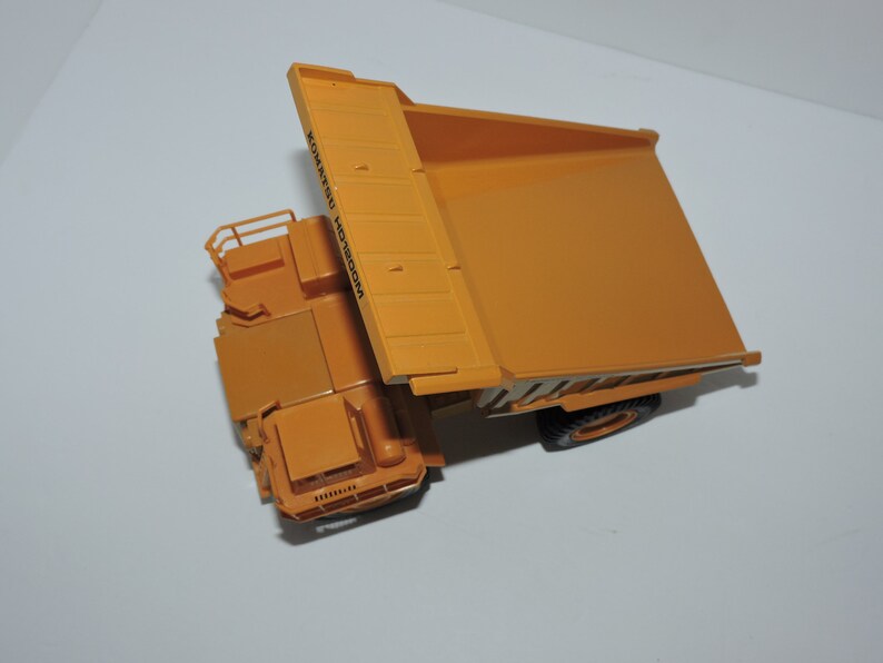 May include: A yellow toy dump truck with the text "KOMATSU" printed on the side. The truck is tilted up, showing the bed of the truck.