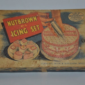 May include: A vintage box for a 'Nutbrown' No. 5 Icing Set. The box features a red and white illustration of a cake with pink icing and a piping bag. The text 'Complete with 6 different nozzles' is printed on the bottom of the box.