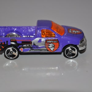 Può includere: Un camioncino di giocattolo viola con un design bianco e arancione. Il camioncino ha la scritta "HWTrans Maintenance Service" sul lato. Il camioncino è un giocattolo Hot Wheels.