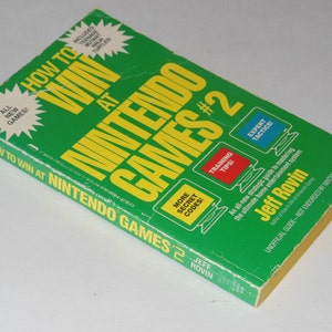 Könnte beinhalten: Grünes und gelbes Buchcover mit dem Titel "How to Win at Nintendo Games #2" von Jeff Rovin. Das Cover zeigt Illustrationen von Spielcontrollern und Text über Trainingstipps, Expertentaktiken und Geheimcodes.