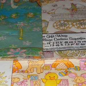 May include: A set of three baby-themed gift wrap rolls. The first roll features a blue and green background with a white stork and flowers. The second roll features a white background with a baby carriage and a small angel. The third roll features a white background with a variety of baby animals and toys. The text on the label reads "Gift Wrap Collage Cadeau Superfin 19" x 27.5" 48 cm x 70 cm Total 7.3 sq. ft. 7.3 pi&sup2; 67 dm&sup2; Card Company, Scarborough, Ont. MIV 2W2 of Bellefair Greeting Card Co. Limited."