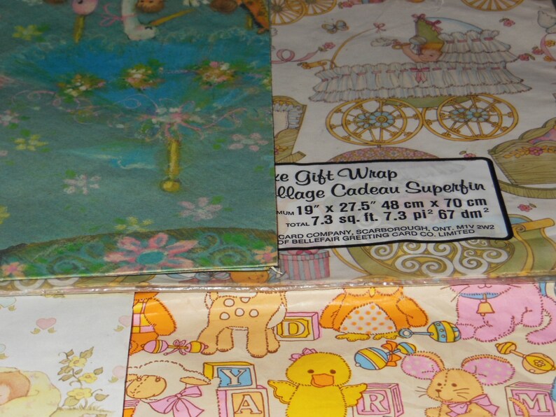 May include: A set of three baby-themed gift wrap rolls. The first roll features a blue and green background with a white stork and flowers. The second roll features a white background with a baby carriage and a small angel. The third roll features a white background with a variety of baby animals and toys. The text on the label reads "Gift Wrap Collage Cadeau Superfin 19" x 27.5" 48 cm x 70 cm Total 7.3 sq. ft. 7.3 pi&sup2; 67 dm&sup2; Card Company, Scarborough, Ont. MIV 2W2 of Bellefair Greeting Card Co. Limited."