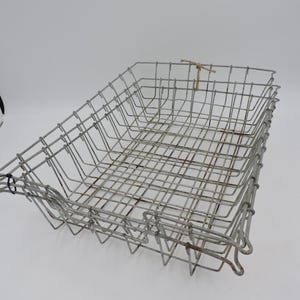 May include: A rectangular metal wire basket with a grid pattern. The basket is made of silver-colored wire and has a slightly rusted appearance. A small piece of twine is tied to the top of the basket. The basket is likely used for storage or display.