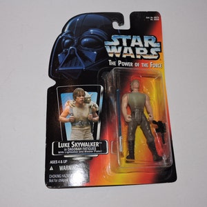 Op de afbeelding: Een Star Wars-actiefiguur van Luke Skywalker in DagoBAH-kleding. De figuur houdt een lightsaber en een blasterpistool vast. De verpakking is geel, oranje en rood met het Star Wars-logo en de woorden "The Power of the Force".