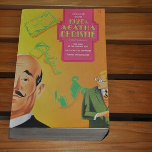 Könnte beinhalten: Ein gelber und rosa Buchumschlag mit dem Titel "1920s Agatha Christie" und dem Text "Band Zwei" oben. Der Umschlag zeigt eine Comic-Illustration eines Mannes mit Schnurrbart und einem Detektiv, sowie einen Schlüssel und einen Geldscheinhaufen.