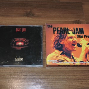 Könnte beinhalten: Zwei Pearl Jam CDs. Die erste CD ist ein schwarzes Case mit einem Foto einer Bühne mit roten Lichtern. Der Text "pearl jam" und "daughter" ist auf der Vorderseite gedruckt. Die zweite CD ist ein gelbes Case mit einem Foto von Eddie Vedder beim Singen. Der Text "the pearl jam Star Profile" ist auf der Vorderseite gedruckt.