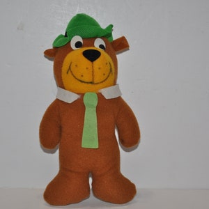 Può includere: Un orso di peluche marrone che indossa un cappello verde e una cravatta verde. L'orso ha un viso giallo con un naso nero e occhi neri.