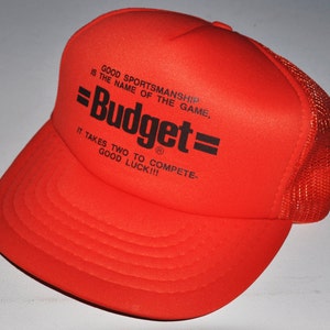 May include: Orange and black mesh trucker hat with the text "GOOD SPORTSMANSHIP IS THE NAME OF THE GAME, =Budget= IT TAKES TWO TO COMPETE- GOOD LUCK!!!"
