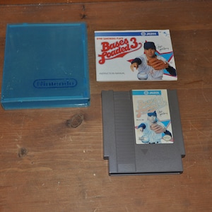 May include: A Nintendo Entertainment System game cartridge and instruction manual for the game "Bases Loaded 3" by Jaleco. The game features a baseball player in a blue and white uniform.