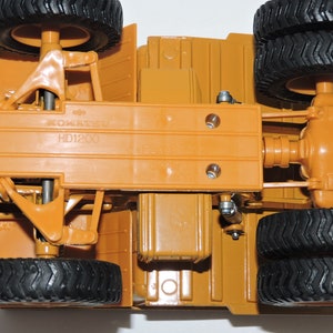 May include: A close-up view of the underside of a toy construction vehicle. The vehicle is brown and has black tires. The vehicle has a label that reads "KOMATSU HD1200".