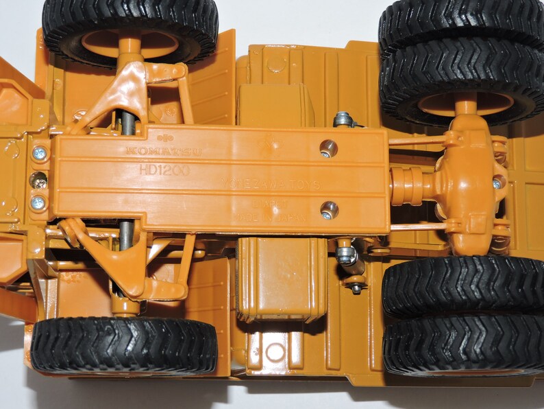 May include: A close-up view of the underside of a toy construction vehicle. The vehicle is brown and has black tires. The vehicle has a label that reads "KOMATSU HD1200".