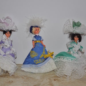 Peut inclure: Trois poupées en porcelaine vintage portant des robes et des chapeaux. Les poupées sont habillées de robes violette, bleue et verte avec des bordures en dentelle blanche. Les poupées portent des chapeaux avec de la dentelle blanche et des rubans. La robe bleue porte l'inscription "Paris" imprimée dessus.