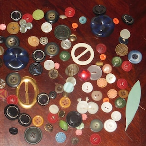 Puede incluir: Una colección de botones vintage en varios colores, formas y tamaños. Los botones están hechos de plástico, metal y madera. Algunos de los botones tienen diseños intrincados, mientras que otros son simples. Los botones están dispuestos en un patrón aleatorio sobre una superficie de madera.