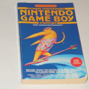 Könnte beinhalten: Ein blaues Buch mit dem Titel "Ultimate Unauthorized Nintendo Game Boy Strategies" von Corey Sandler und Tom Badgett. Das Cover zeigt eine Comic-Katze in einem rosa-gelben Badeanzug, die auf einem Surfbrett auf einer blauen Welle reitet. Das Buch enthält "Inside"-Tipps und Tricks für 40 heiße Spiele, Vorschauen auf 21 zukünftige Spiele und Hintergrundgeschichten für Batman, Spider-Man und andere.