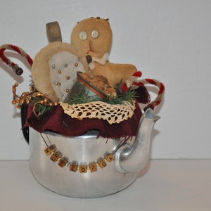 May include: A silver teapot decorated with a gingerbread person, candy canes, and a crocheted doily. The teapot has a chain of letter tiles that spell "HOT COCOA".