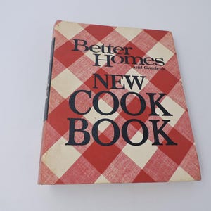 Puede incluir: Un libro de cocina vintage titulado "Better Homes and Gardens New Cook Book". La portada presenta un patrón de cuadros rojos y blancos con el título en letras negras en negrita. El libro de cocina está ligeramente desgastado, lo que sugiere que es un artículo preciado.