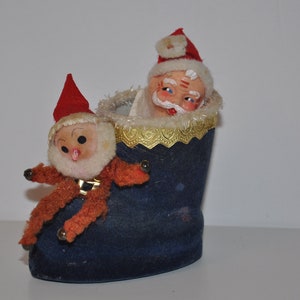May include: A blue felt stocking with a white trim and a gold ribbon. A small Santa Claus doll is peeking out of the top of the stocking, and a smaller elf doll is sitting on the side of the stocking.