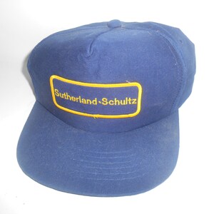 Pode incluir: Um boné de beisebol azul e amarelo com o texto "Sutherland-Schultz" bordado na frente.