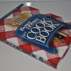 Puede incluir: Un libro de cocina azul con el título "New Better Homes and Gardens Cookbook" sobre un fondo a cuadros rojo y blanco. El libro está abierto y tiene utensilios de cocina en su interior.