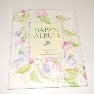 May include: A baby's memory book titled "Baby's Album" in green, with a white cover adorned with a floral design in pink, purple, and green. The book includes the text "A Memory Book With Three-Dimensional Illustrations".