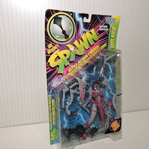 Op de afbeelding: Een Spawn action figure verpakking met een vrouwelijk personage in een zwart en rood kostuum, dat een kettingwapen vasthoudt. De verpakking is helder en kleurrijk met het Spawn-logo en de woorden "Ultra-Action Figures" erop gedrukt.