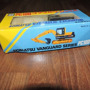 May include: A yellow and black toy Komatsu PC100 hydraulic excavator in a blue and yellow box. The box says "Komatsu Vanguard Series PC100" and "Made in Japan".
