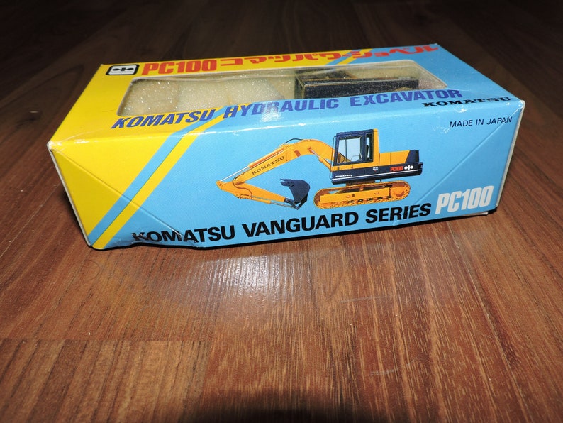 May include: A yellow and black toy Komatsu PC100 hydraulic excavator in a blue and yellow box. The box says "Komatsu Vanguard Series PC100" and "Made in Japan".
