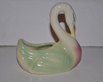 May include: A ceramic swan planter with a green and pink glaze. The swan's neck is curved and its head is tilted down. The planter has a small opening in the back for drainage.