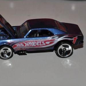 Puede incluir: Un coche de juguete Hot Wheels azul y morado con el capó abierto. El coche tiene gráficos blancos, rojos y azules en el lateral, con las palabras "Hot Wheels" visibles. El número 1 está en el lateral del coche.