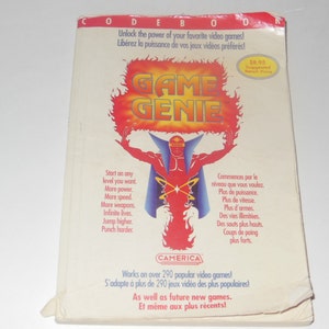 Könnte beinhalten: Eine weiße Box mit einem roten und gelben Game Genie Logo. Die Box hat Text, der "Unlock the power of your favorite video games!" und "Libérez la puissance de vos jeux vidéos préférés!" sagt. Die Box hat auch Text, der "Works on over 290 popular video games!" und "S'adapte à plus de 290 jeux vidéo des plus populaires!" sagt.