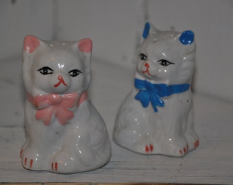 May include: Two white ceramic cat figurines. One cat has a pink bow and the other has a blue bow.