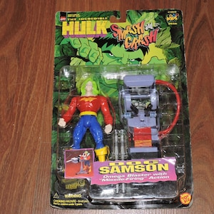 Könnte beinhalten: Eine Plastik-Actionfigur von Doc Samson aus der Zeichentrickserie The Incredible Hulk Smash and Crash. Die Figur trägt ein rot-gelbes Kostüm mit blauer Hose und hat grünes Haar. Die Figur hält eine Waffe und ist in einer Plastik-Blisterverpackung mit einem Hintergrundbild des Hulk verpackt.