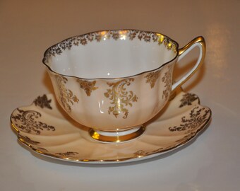 May include: A delicate teacup and saucer set with a pale pink base and intricate gold floral designs. The cup has a scalloped rim and a graceful handle.