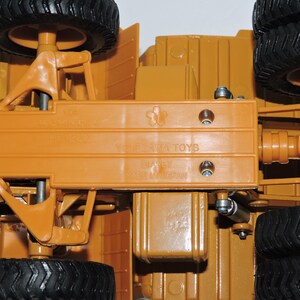 May include: A close-up of the underside of a toy bulldozer. The toy is made of orange plastic and has a metal axle. The toy is labeled "Yonezawa Toys" and "Made in Japan".