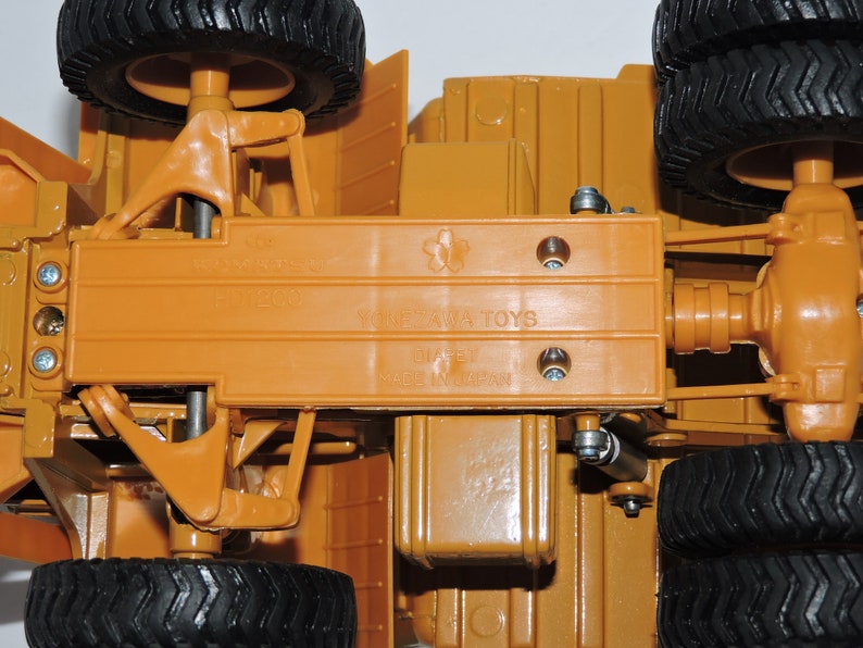 May include: A close-up of the underside of a toy bulldozer. The toy is made of orange plastic and has a metal axle. The toy is labeled "Yonezawa Toys" and "Made in Japan".