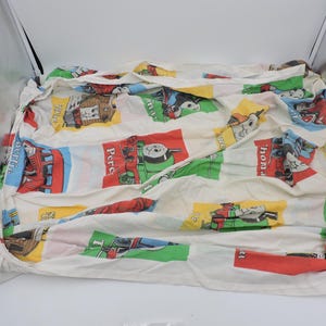 May include: Cream-coloured fitted sheet featuring cartoon train characters in red, green, yellow, and blue blocks. The sheet displays various train designs and character names, offering a playful and vibrant appearance.