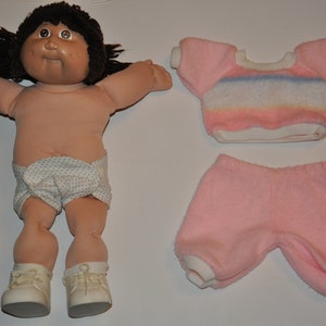 Puede incluir: Una muñeca Cabbage Patch Kid con cabello castaño y un pañal blanco. La muñeca lleva una camisa a rayas rosa y blanca y pantalones cortos rosas.