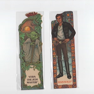 May include: Two Star Wars character bookmark cards. The card on the left features Yoda, the Jedi Master, in a green robe with a walking stick. The card on the right features Han Solo in a black jacket and brown trousers. Both cards have a colourful border.