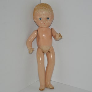 Puede incluir: Una muñeca vintage de plástico duro con la cara pintada y ojos azules. La muñeca tiene un cuerpo rosa claro y le falta algo de pintura. La muñeca está de pie sobre una superficie blanca.