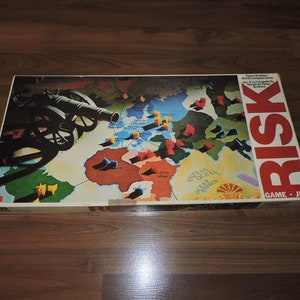 May include: A vintage RISK board game box featuring a world map with territories marked by colored pieces. The box has a red and white "RISK" logo and a cannon illustration. The game is a Parker Brothers World Conquest Game.